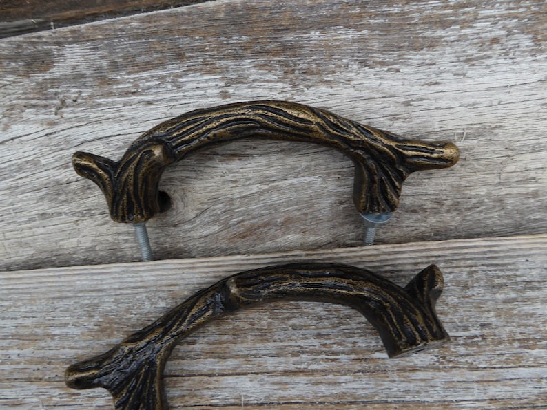 May include: Two bronze-colored, twig-shaped cabinet pulls. The pulls have a textured, bark-like appearance and are mounted with silver screws. The hardware is designed to resemble natural tree branches, adding a rustic touch to furniture.