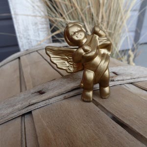 May include: A small, gold-colored cherub figurine with detailed wings. The cherub is standing on a wooden surface, possibly a barrel or similar rustic item. The figurine has a matte finish and is well-lit.