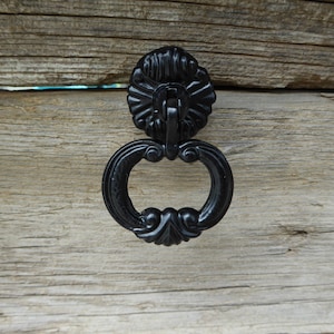 Pode incluir: Puxador de gaveta de metal preto com um design decorativo e ornamentado. O puxador tem a forma de um anel com uma parte superior decorativa.