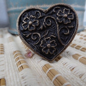 May include: A heart-shaped, antique-style decorative object with a dark bronze finish. The object features an embossed floral design with three flowers and swirling vines. The background is a cream-colored woven surface.