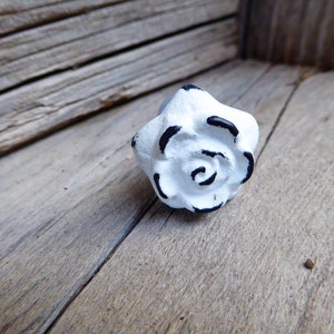 May include: White cast iron rose drawer pull with black detailing. The rose is shown on a weathered wood surface.