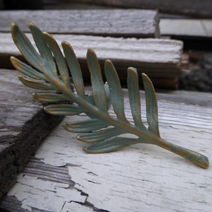 May include: A decorative metal leaf with a blue-green finish and gold accents. The leaf is lying on a weathered, white-painted wooden surface, suggesting a rustic or nature-inspired theme. The leaf's detailed design and color palette create a visually appealing accent piece.