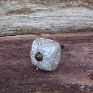 May include: A gray ceramic cabinet knob with a white floral design. The knob has a square shape with a bronze-colored metal base and stem. The knob is on a wooden surface.