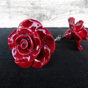May include: Two red ceramic rose drawer pulls with a shiny finish. The rose pulls have a detailed design and are attached to a metal screw.