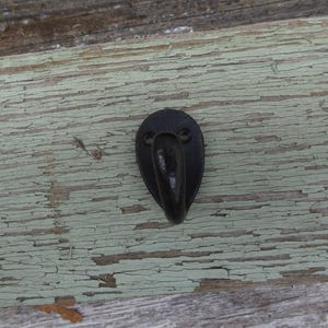 May include: A black, teardrop-shaped metal hook with a curved projection for hanging items. The hook is mounted on a weathered wooden surface with peeling, faded green paint, creating a rustic aesthetic.