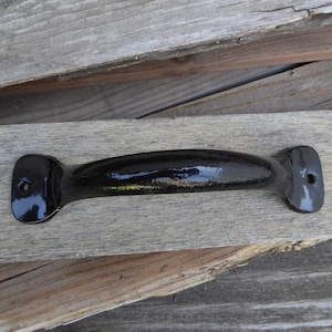 Peut inclure: Poignée de porte en métal noir avec une surface lisse légèrement incurvée. La poignée est présentée sur un fond en bois.