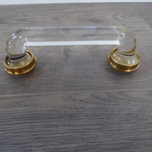 May include: A clear glass cabinet pull with gold-colored metal accents. The handle is curved and has a rectangular shape. The gold accents are circular and provide a decorative base for the glass handle. The hardware is designed for furniture.