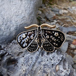 Op de afbeelding: Een zwart en goudkleurige emaille pin in de vorm van een mot. De pin heeft ingewikkelde details, waaronder bloemen en geometrische patronen op de vleugels. De pin heeft een goudkleurige metalen omtrek en een gouden balk aan de bovenkant.