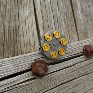 May include: A silver-colored bullet cylinder with six gold-colored bullet casings, each marked "USA AUTO". The cylinder is set against a weathered wooden surface with rusty metal hardware.