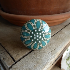 May include: A round ceramic knob with a floral design. The knob features a teal and white flower pattern with a dark green center. It is set against a weathered wooden surface, with a terracotta pot in the background.