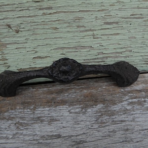 May include: A black, ornate drawer pull with a central decorative element and curved handles. The hardware is made of cast iron and has a textured surface. The drawer pull is designed for use on cabinets or furniture.