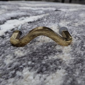 May include: A gold-colored, curved cabinet pull with screws attached. The hardware has a wavy design and is made of a shiny, metallic material. The pull is designed for use on drawers or cabinets.