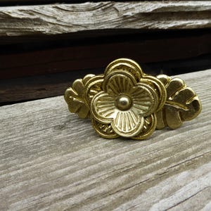 May include: A gold-colored, floral-shaped decorative object. The object features a central flower design with detailed petals and a small, round center. Two leaf-shaped elements flank the flower. The object is placed on a weathered wooden surface.