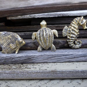 May include: Three metallic gold-toned decorative figures shaped like sea creatures, including an angelfish, a sea turtle, and a seahorse. Each piece features a textured, ridged surface with dark antiqued detailing, showcasing distinct marine life forms.