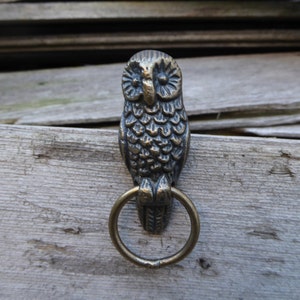 May include: A bronze owl-shaped hook with a ring attached to the bottom. The owl is perched on a wooden surface.