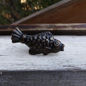 May include: A bronze-colored fish-shaped cabinet knob. The knob has a detailed scale pattern and a textured finish. The fish is positioned on a weathered white wooden surface, suggesting a rustic or nautical theme.