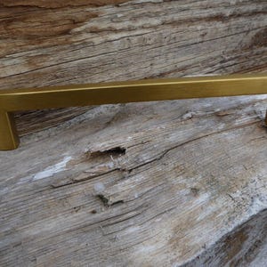 May include: A gold-colored, rectangular cabinet pull with a square, modern design. The hardware is made of metal and is set against a weathered, light brown wooden surface. The pull is approximately 6 inches long.