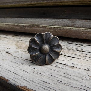 May include: A bronze-coloured, flower-shaped cabinet knob. The knob features a detailed petal design and a central, rounded core. It sits on a weathered, white-painted wooden surface, with a backdrop of wooden planks.