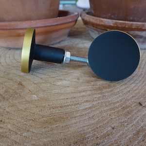Puede incluir: Pomo redondo negro y dorado para armario con tornillo de metal. El pomo tiene una cara plana circular y una base redonda.