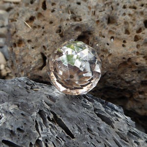 May include: A clear, faceted crystal ball rests on a dark gray, textured rock. The crystal reflects the surrounding environment, creating a sparkling effect. The background features a larger, porous, brown rock.