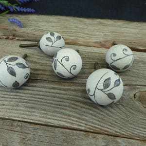 May include: A set of four round, off-white ceramic cabinet knobs, each featuring a hand-painted dark grey vine and leaf pattern. The knobs have a smooth, spherical shape and include a metal screw attachment extending from the back of each piece.