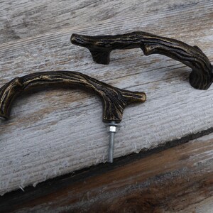May include: Two bronze-colored cabinet pulls in the shape of tree branches. Each pull has a curved handle with a textured surface, mounted with a bolt and nut. The pulls are set against a weathered wooden background.