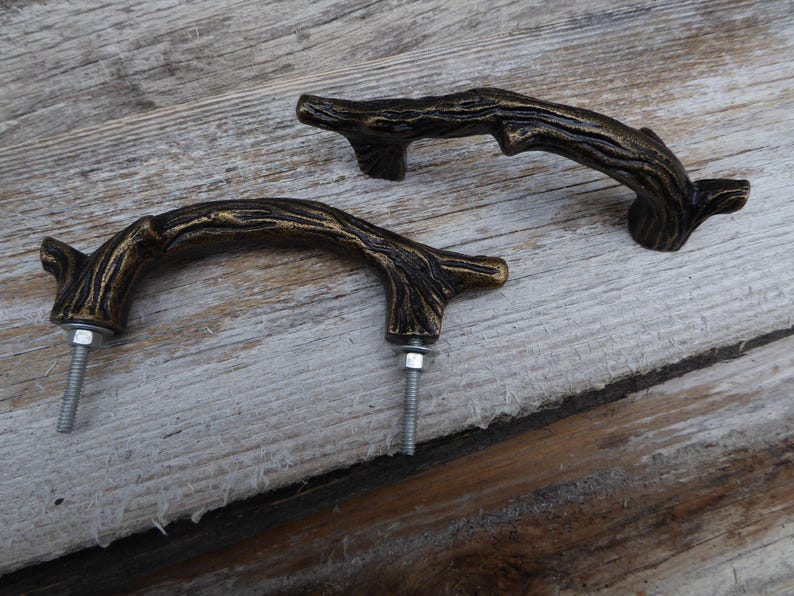 May include: Two bronze-colored cabinet pulls in the shape of tree branches. Each pull has a curved handle with a textured surface, mounted with a bolt and nut. The pulls are set against a weathered wooden background.