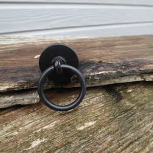 May include: Black metal ring pull handle with a round base. The handle is attached to a piece of weathered wood.