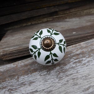 May include: A white ceramic cabinet knob with a green leaf design and a brass-colored center. The knob is round and sits on a weathered wooden surface. Decorative hardware for furniture.