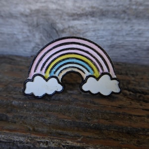 May include: A rainbow-colored enamel pin with white clouds on either side. The rainbow has pink, yellow, blue, and white stripes.