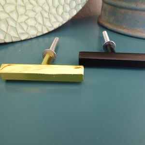 Può includere: Due pomelli per armadio, uno dorato e uno nero, con un design a forma di T. I pomelli sono su una superficie blu.