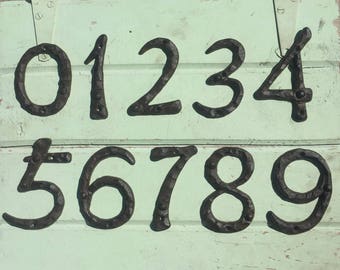 May include: Black metal house numbers 0 through 9 on a light green wooden surface. The numbers are in a rustic style with a hammered finish.