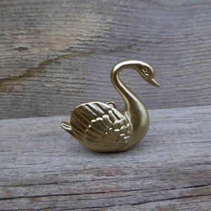 May include: A golden swan figurine with detailed wing and feather textures. The swan has a curved neck and is positioned on a weathered wooden surface. The figurine is a decorative item.