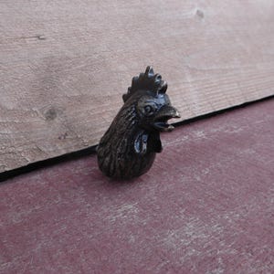 Puede incluir: Un llamador de puerta de hierro fundido negro con un diseño detallado. El gallo está mirando hacia la derecha con el pico abierto.