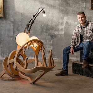 May include: A wooden sculpture of a fish with a large mouth and two light bulbs. The sculpture is lit up and is sitting on a wooden base. A man is sitting on a wooden platform with a large metal number 3 behind him.