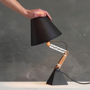 Peut inclure: Lampe de bureau noire avec abat-jour conique, bras en bois et m&eacute;tal, et base triangulaire. La lampe est r&eacute;glable et poss&egrave;de un cordon d'alimentation noir. La lampe mesure environ 38 cm de haut.