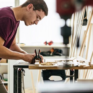Peut inclure: Une personne travaillant sur un projet de menuiserie, utilisant des pinces pour fixer des morceaux de bois. L'atelier comprend un &eacute;tabli, diverses pi&egrave;ces de bois et des outils, sugg&eacute;rant un travail de menuiserie ou d'artisanat.