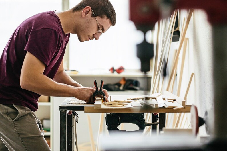 Peut inclure: Une personne travaillant sur un projet de menuiserie, utilisant des pinces pour fixer des morceaux de bois. L'atelier comprend un &eacute;tabli, diverses pi&egrave;ces de bois et des outils, sugg&eacute;rant un travail de menuiserie ou d'artisanat.