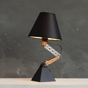 Peut inclure: Une lampe de bureau noire avec un abat-jour conique. La lampe est dot&eacute;e d'un bras articul&eacute; en bois et en m&eacute;tal et d'une base g&eacute;om&eacute;trique noire. L'abat-jour de la lampe est noir et l'int&eacute;rieur est dor&eacute;. La lampe est sur une surface blanche contre un mur gris fonc&eacute;.