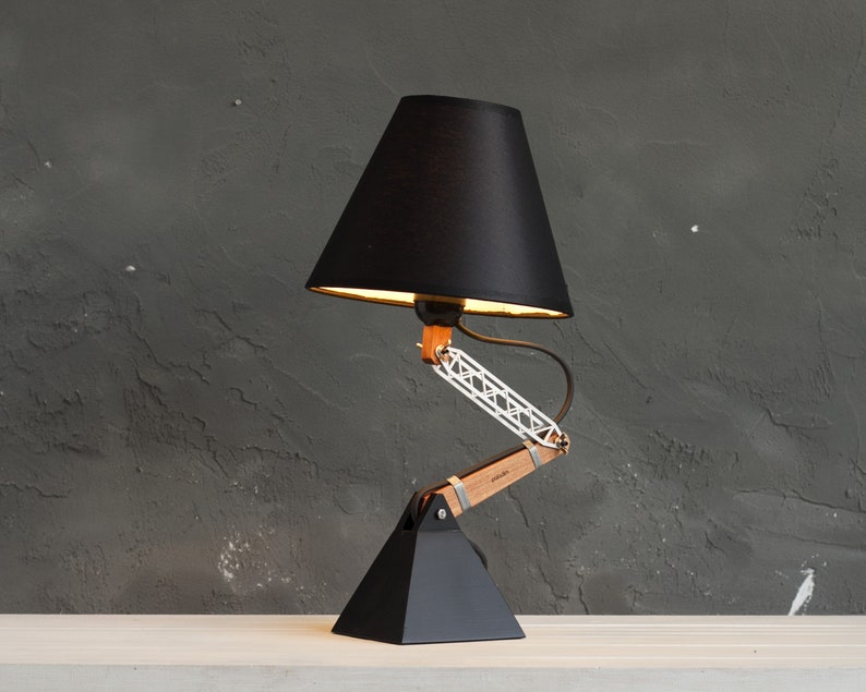 Peut inclure: Une lampe de bureau noire avec un abat-jour conique. La lampe est dot&eacute;e d'un bras articul&eacute; en bois et en m&eacute;tal et d'une base g&eacute;om&eacute;trique noire. L'abat-jour de la lampe est noir et l'int&eacute;rieur est dor&eacute;. La lampe est sur une surface blanche contre un mur gris fonc&eacute;.