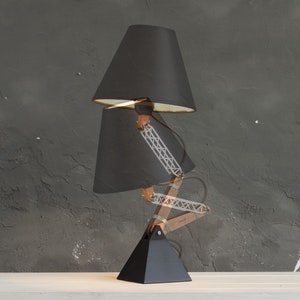 Peut inclure: Lampe de bureau noire avec un design &agrave; double abat-jour. La lampe a une base noire, des bras en bois et une structure de support en m&eacute;tal. Les abat-jours sont noirs avec des int&eacute;rieurs dor&eacute;s. La lampe est sur une surface blanche.