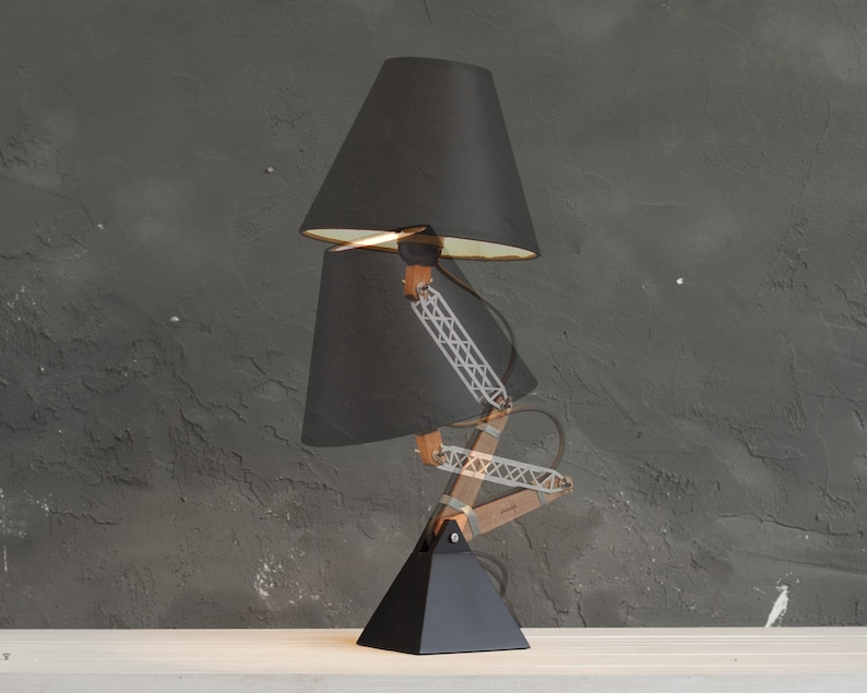 Peut inclure: Lampe de bureau noire avec un design &agrave; double abat-jour. La lampe a une base noire, des bras en bois et une structure de support en m&eacute;tal. Les abat-jours sont noirs avec des int&eacute;rieurs dor&eacute;s. La lampe est sur une surface blanche.