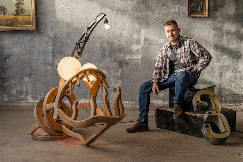 May include: A wooden sculpture of a fish with a large mouth and two light bulbs. The sculpture is lit up and is sitting on a wooden base. A man is sitting on a wooden platform with a large metal number 3 behind him.