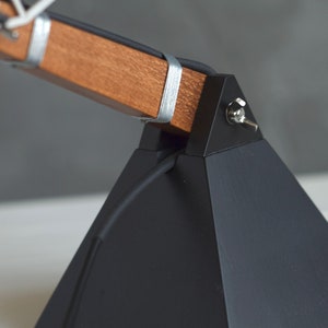 Peut inclure: Gros plan sur une lampe de bureau moderne avec une base g&eacute;om&eacute;trique noire et un bras en bois. Le bras est fix&eacute; avec des bandes m&eacute;talliques argent&eacute;es. Un petit interrupteur est visible sur le c&ocirc;t&eacute; de la base. Design minimaliste et industriel.
