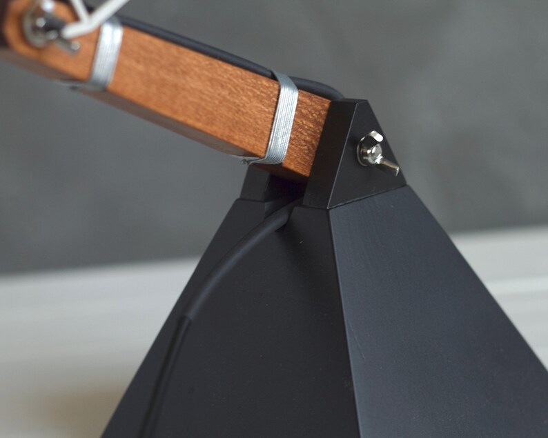 Peut inclure: Gros plan sur une lampe de bureau moderne avec une base g&eacute;om&eacute;trique noire et un bras en bois. Le bras est fix&eacute; avec des bandes m&eacute;talliques argent&eacute;es. Un petit interrupteur est visible sur le c&ocirc;t&eacute; de la base. Design minimaliste et industriel.
