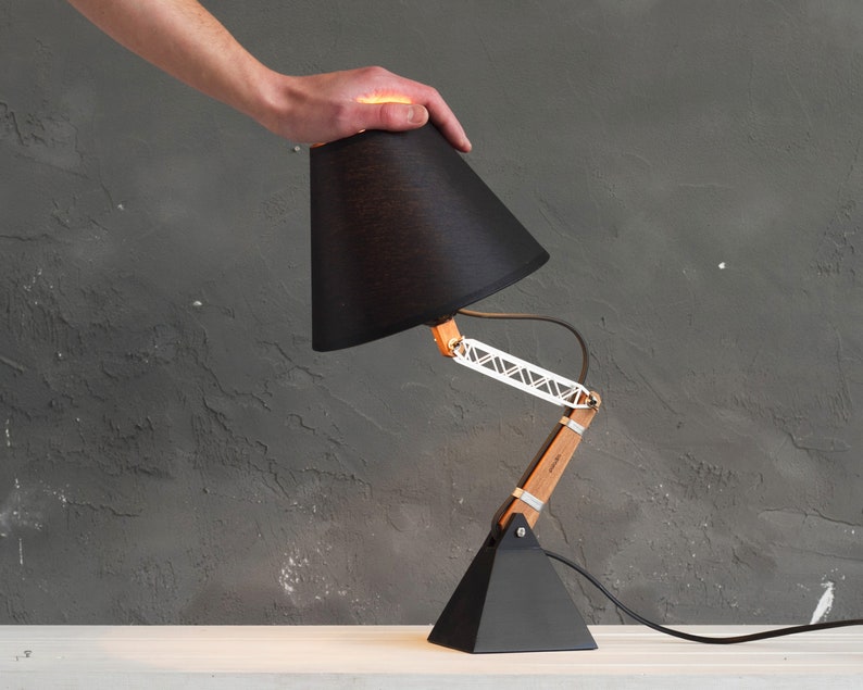 Peut inclure: Lampe de bureau noire avec abat-jour conique, bras en bois et m&eacute;tal, et base triangulaire. La lampe est r&eacute;glable et poss&egrave;de un cordon d'alimentation noir. La lampe mesure environ 38 cm de haut.