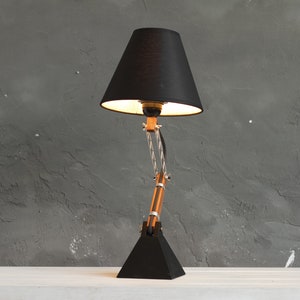 Peut inclure: Une lampe de bureau noire avec un abat-jour conique et un bras r&eacute;glable. La lampe a une base noire et un bras en bois et en m&eacute;tal. L'abat-jour est noir et la lumi&egrave;re est allum&eacute;e. La lampe est sur une surface blanche contre un mur gris.