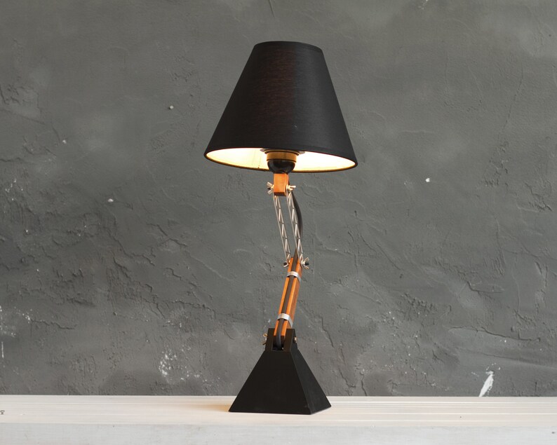 Peut inclure: Une lampe de bureau noire avec un abat-jour conique et un bras r&eacute;glable. La lampe a une base noire et un bras en bois et en m&eacute;tal. L'abat-jour est noir et la lumi&egrave;re est allum&eacute;e. La lampe est sur une surface blanche contre un mur gris.