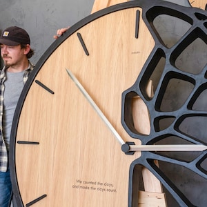 May include: Large wooden clock with a light wood face and black frame. The clock features black stick hour markers and silver hands. The right side of the clock has a unique open design. The clock has the text "We counted the days, and made the days count."