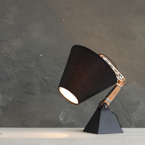 Peut inclure: Une lampe de bureau noire moderne avec un abat-jour conique. La lampe est dot&eacute;e d'un bras en bois et d'une structure m&eacute;tallique g&eacute;om&eacute;trique. La base est de forme pyramidale noire. La lampe est sur une surface blanche contre un mur gris textur&eacute;.